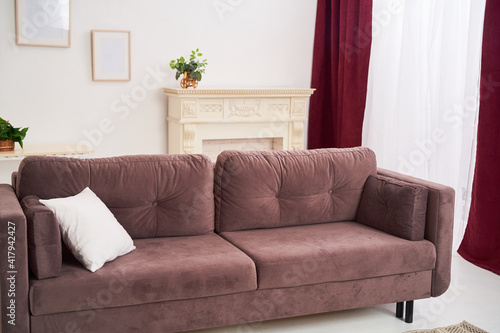 bright, cozy home room with a large brown sofa, a modern wicker carpet, a mat and a fireplace with large interior candles. The large window is framed with tulle and curtains, blackout curtains. © skif