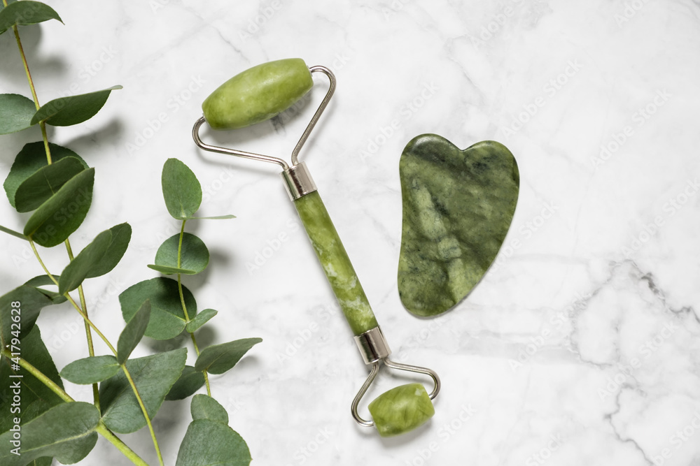 Obraz na płótnie Green jade roller and gua sha stone for facial massage and eucalyptus branch on marble background. Home beauty and selfcare accessories. Face roller for anti age wrinkle treatment. Top view, flat lay. w salonie