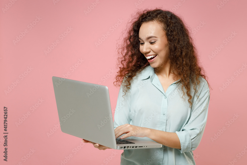 Young black african american smiling fun freelancer curly woman 20s wear blue shirt hold using laptop pc computer chatting browsing internet isolated on pastel pink color background studio portrait Young black african american smiling fun freelancer curly woman 20s wear blue shirt hold using laptop pc computer chatting browsing internet isolated on pastel pink color background studio portrait