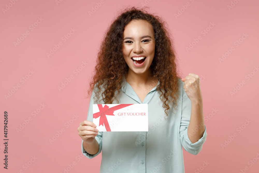 Young black african excited cute rich curly woman 20s wear blue shirt holding gift voucher flyer mock up do winner gesture clench fist celebrating isolated on pastel pink background studio portrait Young black african excited cute rich curly woman 20s wear blue shirt holding gift voucher flyer mock up do winner gesture clench fist celebrating isolated on pastel pink background studio portrait