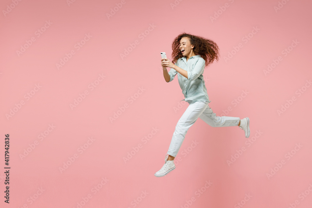 Full length side view young black african fun happy smiling curly student woman 20s in blue shirt holding mobile cell phone chatting jump high run isolated on pastel pink background studio portrait.