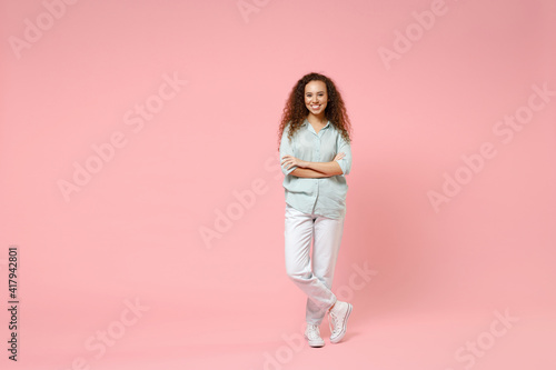 Full length young black african american happy smiling positive curly woman 20s wearing blue casual shirt looking camera hold hands crossed folded isolated on pastel pink background studio portrait.