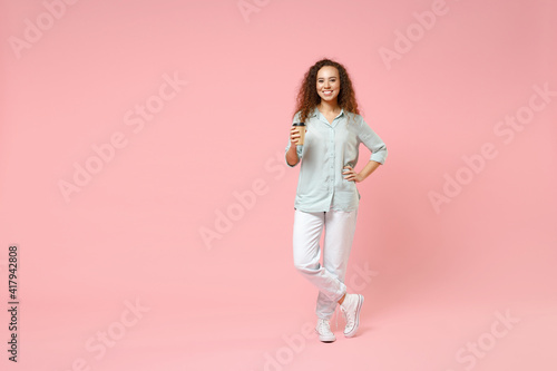 Full length young black african fun happy smiling stylish curly student woman 20s in blue shirt holding paper cup of coffe arm akimbo on waist isolated on pastel pink color background studio portrait.