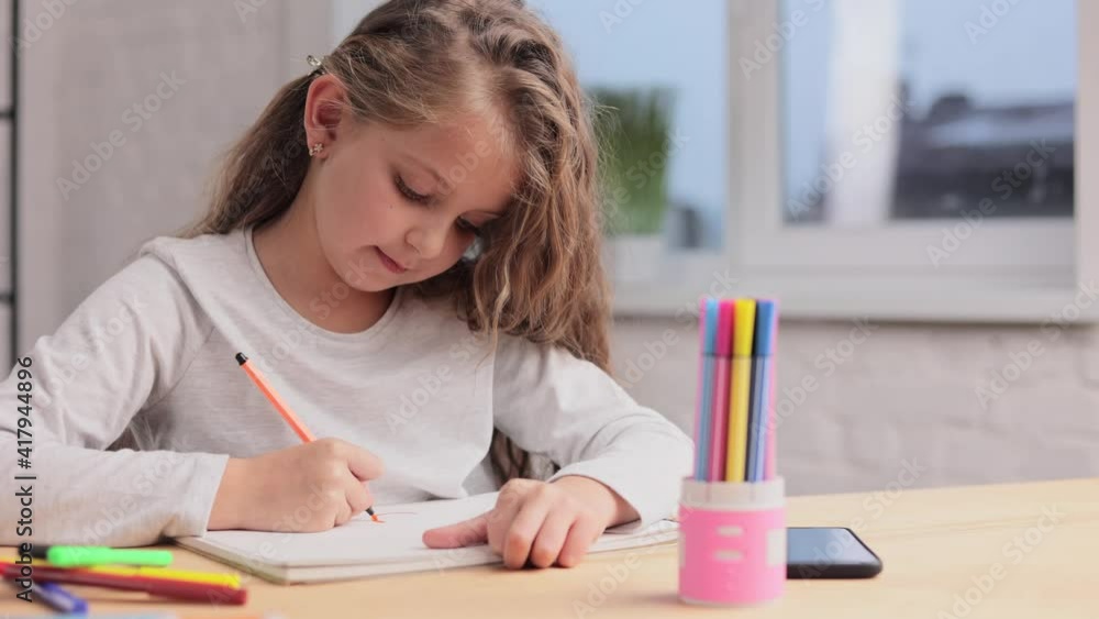 Cute little girl are drawing pictures with felt tip pen on the white paper album sitting at the table. Playing alone, creative art activity at home. Spending free time at home during school holidays Cute little girl are drawing pictures with felt tip pen on the white paper album sitting at the table. Playing alone, creative art activity at home. Spending free time at home during school holidays
