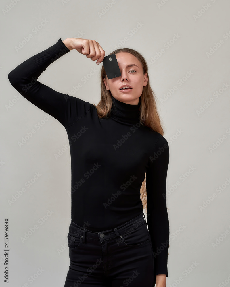 Pretty young woman wearing black clothes looking at camera, holding plastic credit card over her eye while posing isolated over gray background Pretty young woman wearing black clothes looking at camera, holding plastic credit card over her eye while posing isolated over gray background