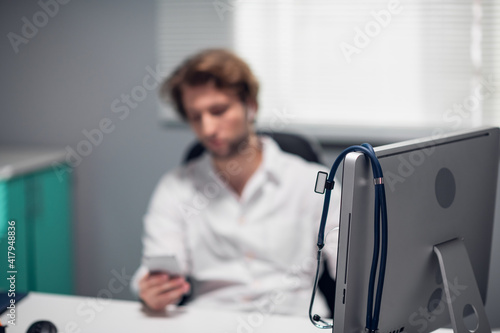 Blurred picture of a doctor sitting at his desk in the office, a fancy monitor in the foreground with a stethoscope hanging on it.