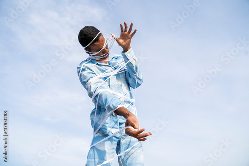 Young man wearing cloud patterned denim outfit struggling to get free from rope while standing against sky