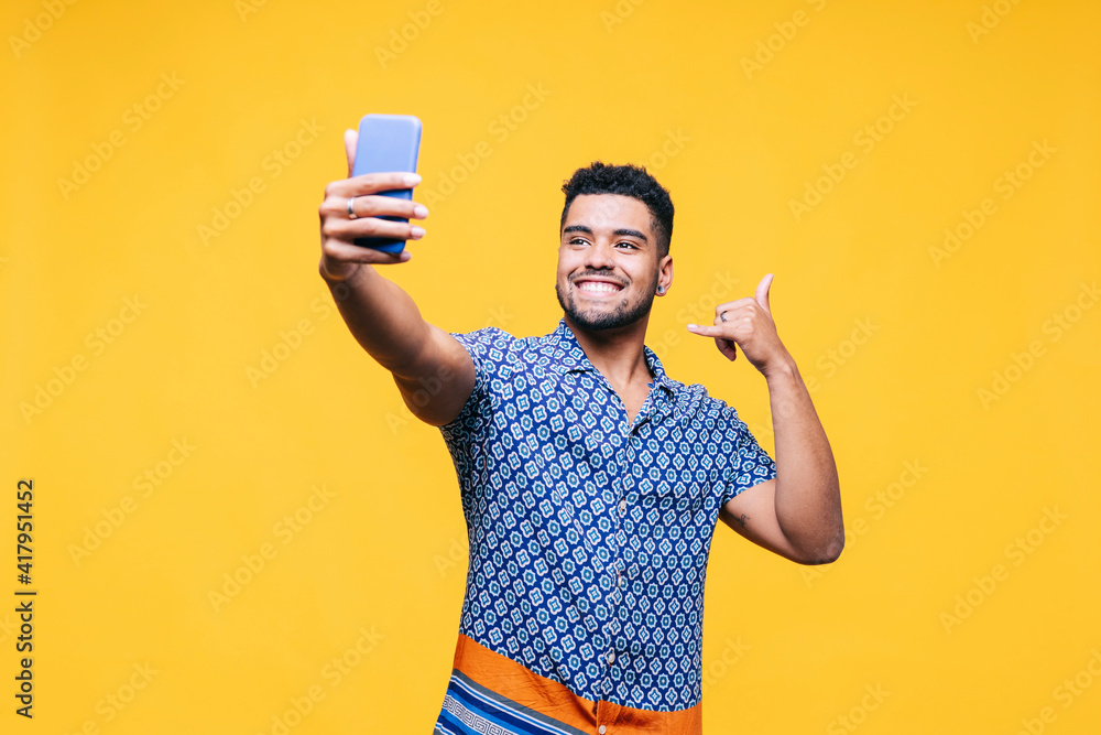Smiling man gesturing while taking selfie through mobile phone against yellow background Smiling man gesturing while taking selfie through mobile phone against yellow background