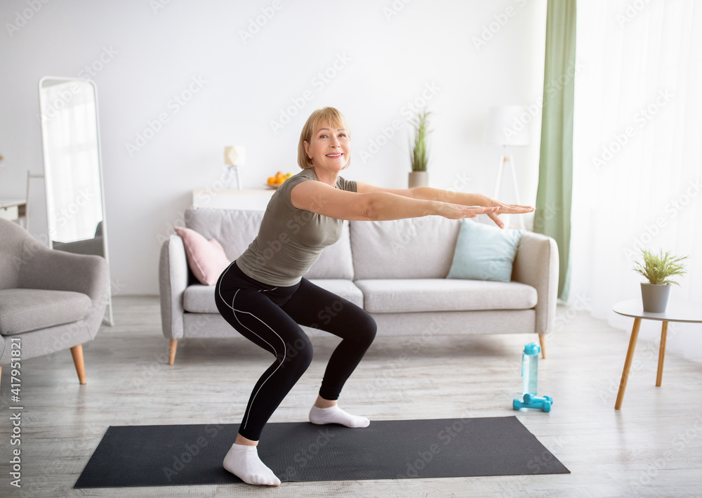 Home workout concept. Beautiful mature lady in sportswear doing squats on yoga mat indoors, free space