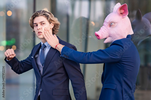Young male entrepreneur wearing pig mask slapping colleague by glass wall photo