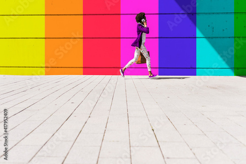 Young woman talking on smart phone while walking by colorful wall during sunny day