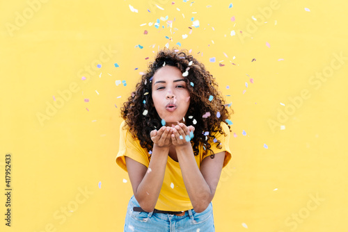 Young woman blowing multi colored confetti against yellow wall