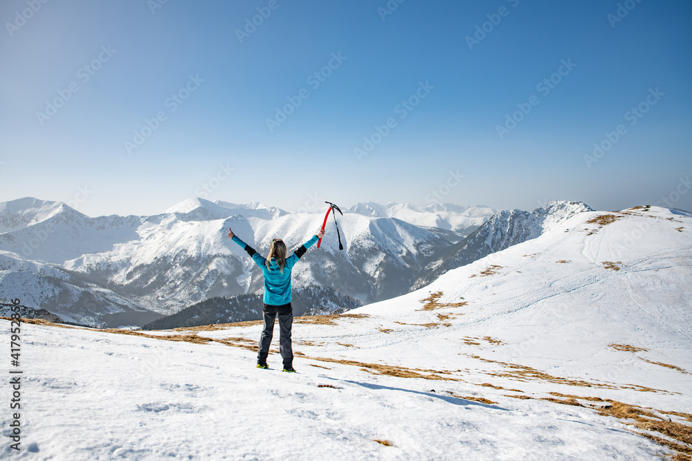 Female tourist with raised hands on winter trail in mountains. A tourist is holding an ice ax in her hand.