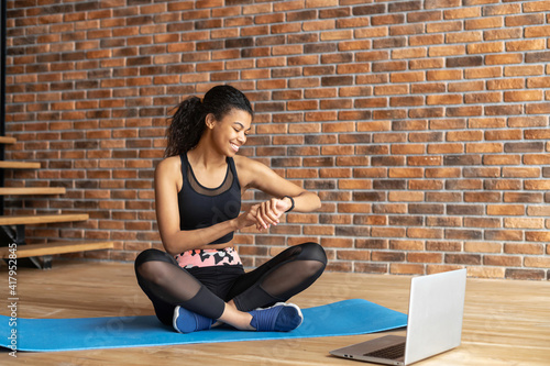 Charming young African-American woman doing sports workout at home with a laptop, a multiracial girl controls pulse rate and calorie loss with a fitness tracker band