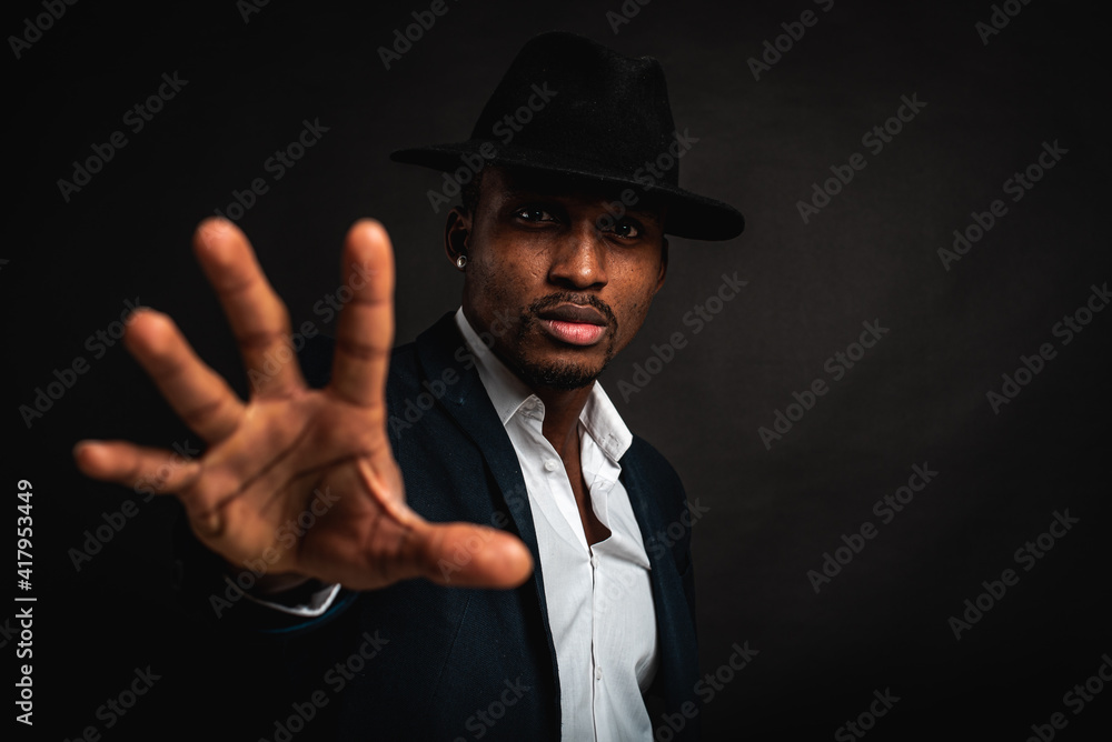 Young man in African American suit standing in front of camera with hand in front as stop sign, on dark background. Selective focus. Sign language.
