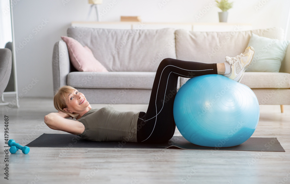Fit mature lady doing abs exercises on yoga mat at home, training her core muscles with fitball, copy space