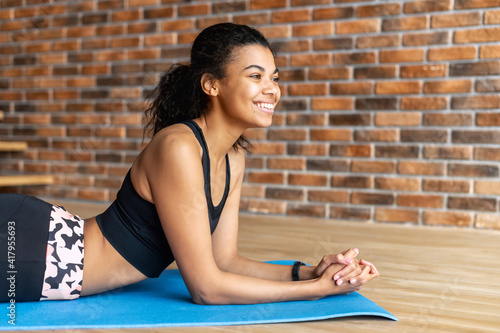 A portrait of a fit African-American woman in sports outfit laying on the mat and looks away a smile, takes a break of training. Sports at home concept