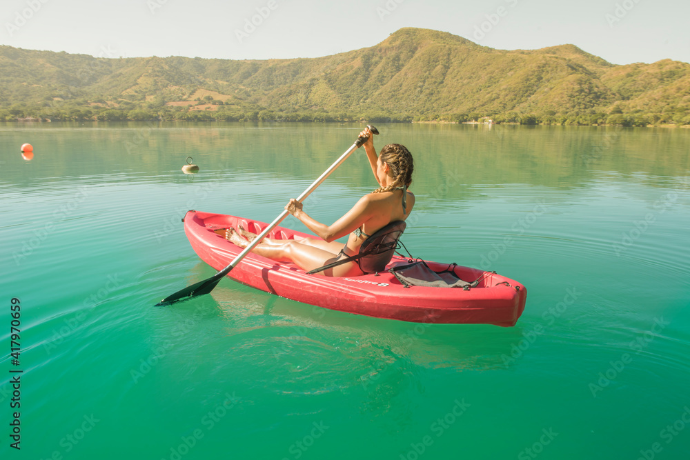 Latin woman paddling stand up on paddle, she is enjoying her vacation, she is in a red kayak on a turquoise lake and mountains in the background, behind her a summer sunset