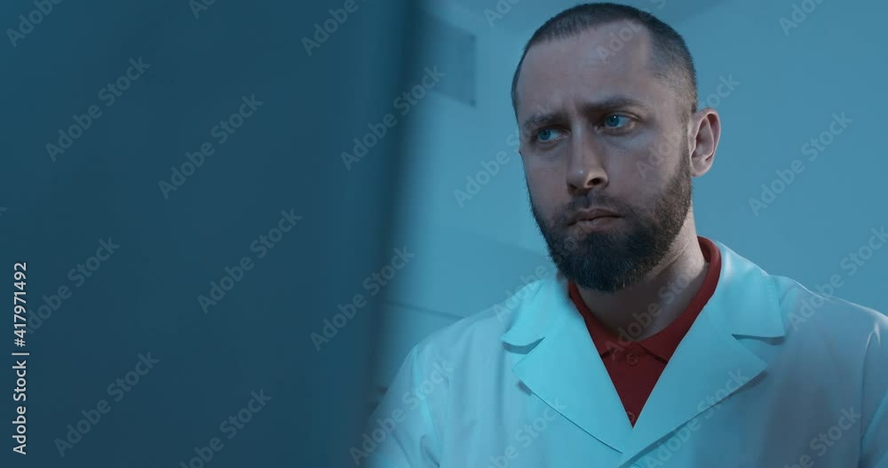 A male medical worker is sitting in a dark room at the desk in front of a computer; he is looking at the screen and looks serious.