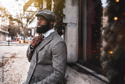 The portrait of a fashionable mature bearded black guy in an elegant suit and a hat leaning on the wall with a window decorated with needles of an artificial Christmas tree with a garland