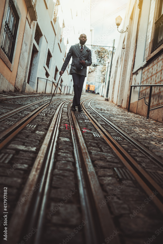 A dapper bald bearded guy in an elegant custom-made business costume and eyeglasses is leaning on his walking stick while staying in the middle of a narrow antique European street on the tramway track