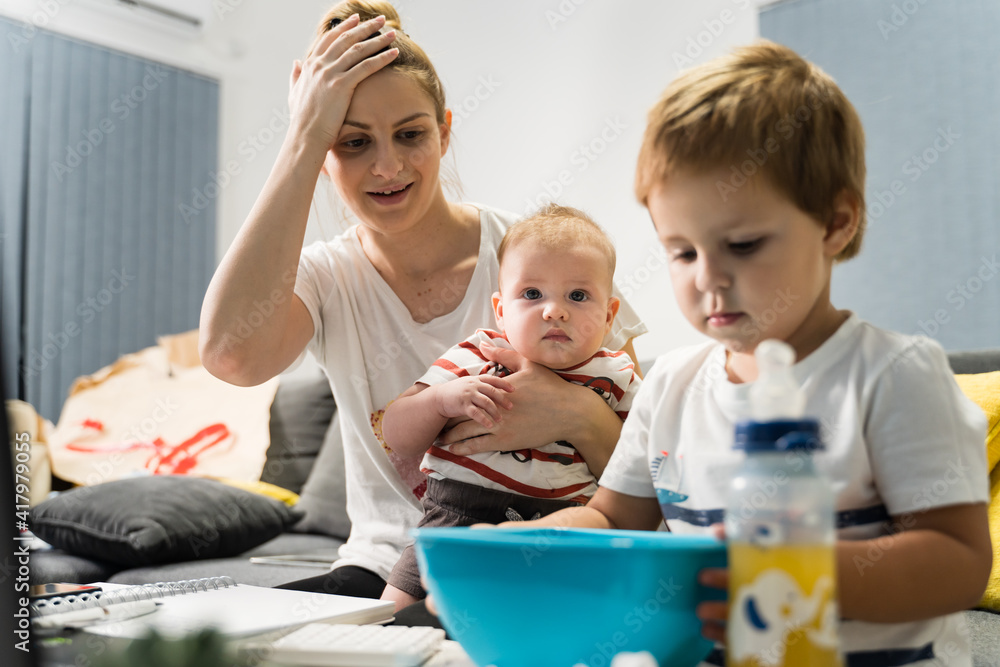 Small caucasian boy eating snacks at home with his mother and baby brother or sister behind watching - little kid appetite for candy and food growing up development concept Small caucasian boy eating snacks at home with his mother and baby brother or sister behind watching - little kid appetite for candy and food growing up development concept