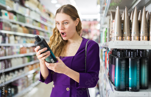 Attractive girl standing near shelves with haircare cosmetics in supermarket, looking in amazement at shampoo bottle