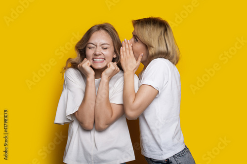 Two caucasian women are gossiping on a yellow studio wall smiling while wearing white t-shirt