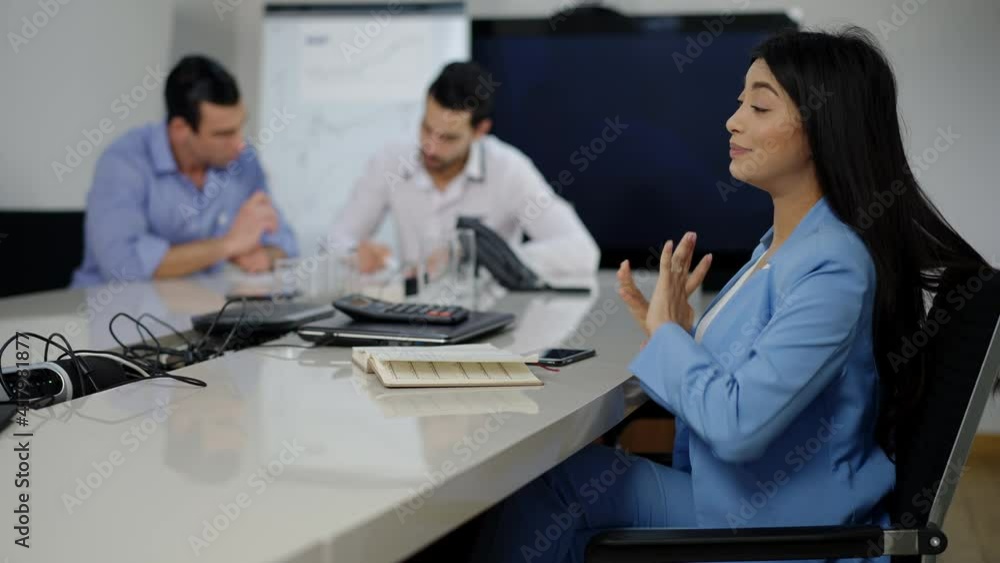 Young slim Asian woman stretching sitting in office as blurred Middle Eastern men talking at background. Side view portrait of exhausted millennial overworking indoors. Lifestyle and business. Young slim Asian woman stretching sitting in office as blurred Middle Eastern men talking at background. Side view portrait of exhausted millennial overworking indoors. Lifestyle and business.