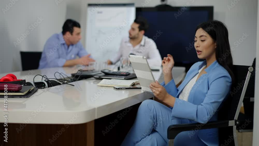 Confident beautiful Asian businesswoman sitting at conference table using tablet with blurred Middle Eastern men talking at background. Portrait of successful young millennial in office indoors. Confident beautiful Asian businesswoman sitting at conference table using tablet with blurred Middle Eastern men talking at background. Portrait of successful young millennial in office indoors.
