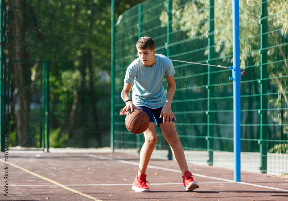 Cute Teenager in green t-shirt with orange basketball ball plays basketball on street playground in summer. Hobby, active lifestyle, sports activity for kids.	