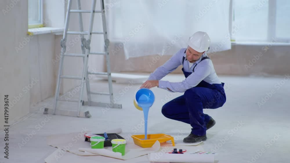 portrait of young smiling man in work clothes and cap pours blue dyestuff in paint tray for painting walls during renovation, smiling and looking at camera portrait of young smiling man in work clothes and cap pours blue dyestuff in paint tray for painting walls during renovation, smiling and looking at camera
