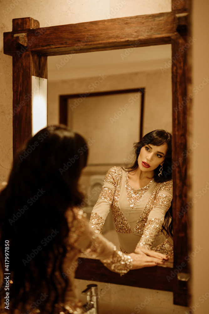 Beauty glamour brunette lady with bright make up  looking in the mirror. Beautiful gorgeous woman in beautiful evening dress in Luxurious style room. Elegant glamorous lady portrait in golden dress