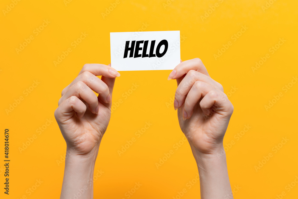 Woman's hands hold a white card with the inscription Hello. Close up. Yellow background. The concept of the world hello day Woman's hands hold a white card with the inscription Hello. Close up. Yellow background. The concept of the world hello day