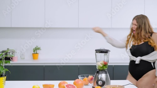 Dieting and nutrition concept. Dancing happy overweight girl dressed in black leotard and white shirt. Dancing in the kitchen curvy body girl blondie preparing fresh fruits juices. FHD footage.  photo