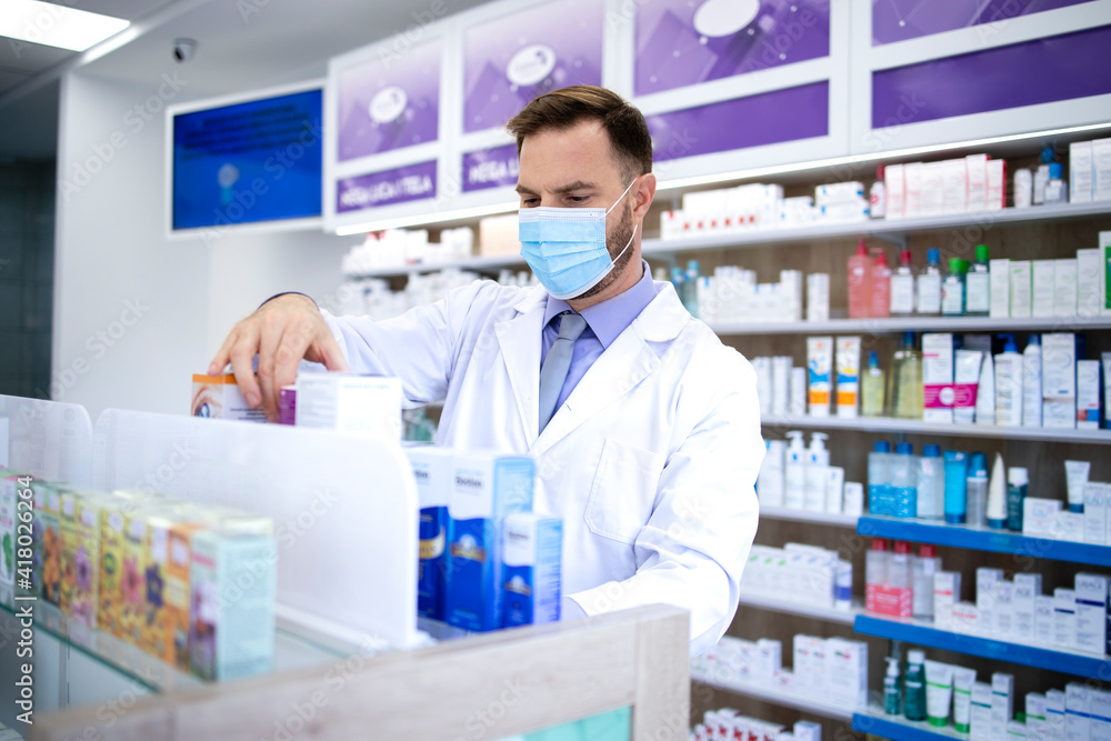 Pharmacist wearing face mask and white coat working in pharmacy store during corona virus pandemic arranging medicines on the shelf. Healthcare and medicine.
