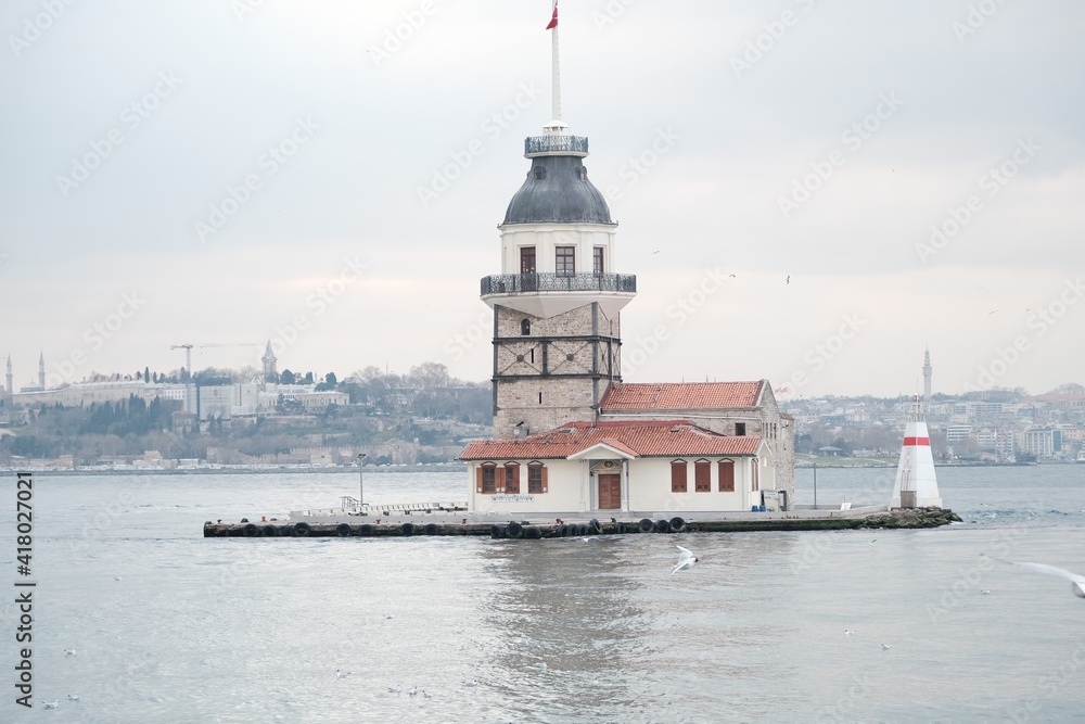 The Maiden's Tower under gray cloudy sky, Bosphorus, Istanbul, Turkey during overcast weather with sunshine reflection in bosporus sea.  Groups of seagulls flying on sea. İstanbul Turkey 01.03.2021