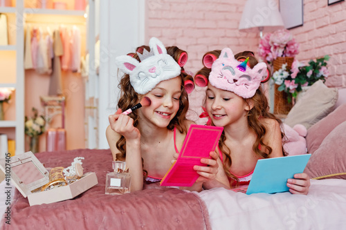 Children's pajama party. A teenage girl in pink pajamas, with makeup and pink curlers on the bed with makeup brushes is making up.