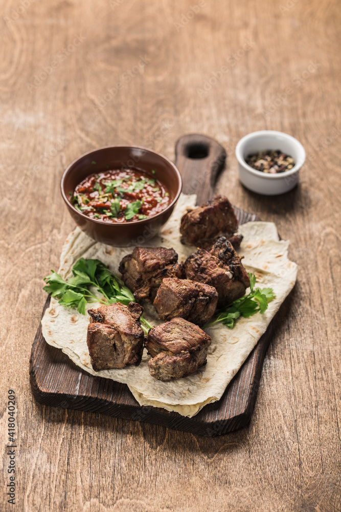Pork shish kebab on a wooden board with tomato sauce on a dark background. Grilled picnic food. Side view with a copy space for the text. Pork shish kebab on a wooden board with tomato sauce on a dark background. Grilled picnic food. Side view with a copy space for the text.