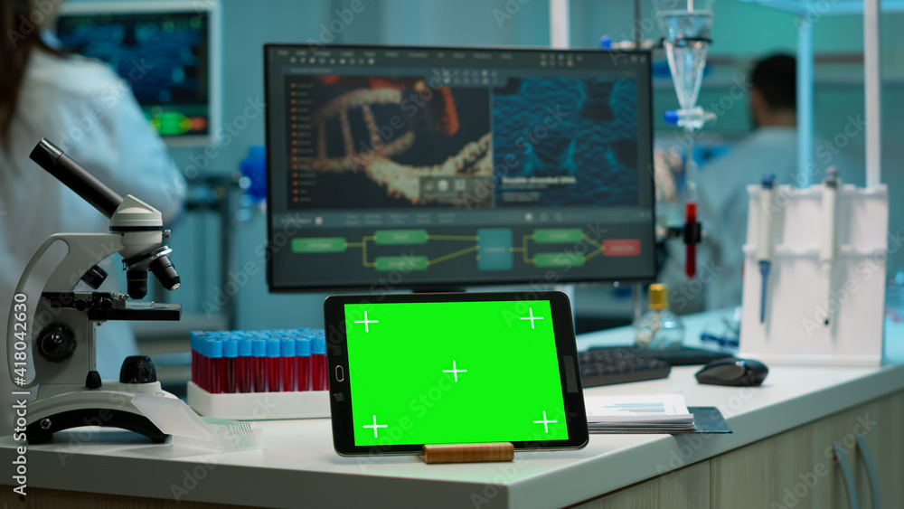 Display tablet with green screen, mock up on template placed on desk in scientific laboratory while woman medical research scientist analysing virus evolution at digital monitor conducting experiment