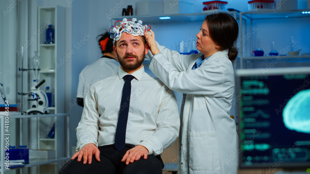 Woman researcher putting brainwave scanning headset to patient developing experiments using brain study lab monitors. Man siting on chair while scientist adjusting device examining nervous system Woman researcher putting brainwave scanning headset to patient developing experiments using brain study lab monitors. Man siting on chair while scientist adjusting device examining nervous system