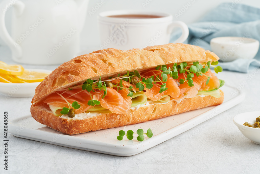 Big sandwich with salmon, cream cheese, cucumber slices on white concrete table. Morning healthy breakfast with fish and cup of drink, side view Big sandwich with salmon, cream cheese, cucumber slices on white concrete table. Morning healthy breakfast with fish and cup of drink, side view