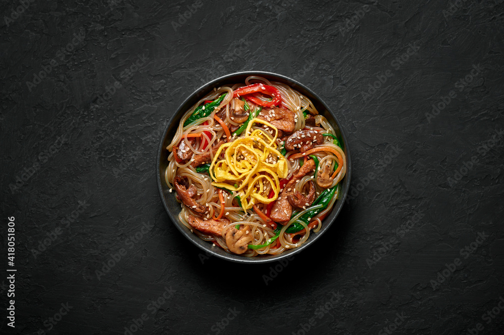 Japchae in black bowl on dark slate table top. Korean cuisine glass chapchae noodles dish with vegetables and meat. Asian traditional food. Authentic meal. Top view