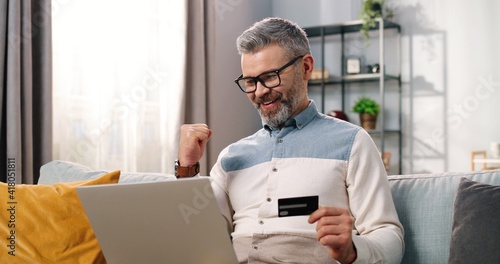Close up of Caucasian middle-aged bearded man in glasses sitting on sofa in cozy room tapping on laptop computer using credit card buying online in good mood satisfied making YES gesture, e-commerce photo