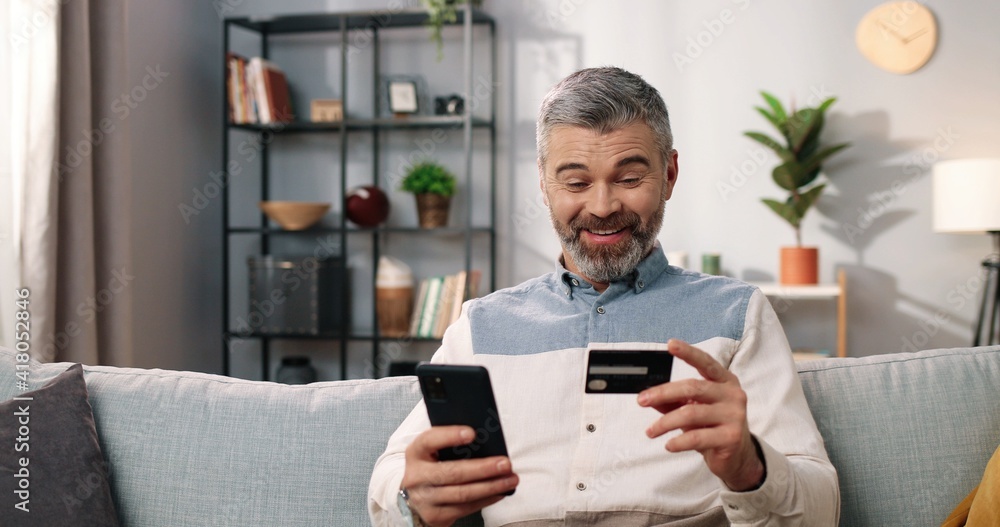 Close up of joyful Caucasian middle-aged bearded man sitting on couch in cozy room tapping on smartphone using credit card buying online in good mood satisfied making YES gesture, e-commerce, shopping Close up of joyful Caucasian middle-aged bearded man sitting on couch in cozy room tapping on smartphone using credit card buying online in good mood satisfied making YES gesture, e-commerce, shopping