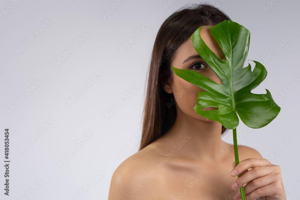 Handsome caucasian woman. Spa and Beauty concept. Closed female face by monstera leave. Spa salon or organic cosmetics logotype. Woman Face on gray background with side space. Handsome caucasian woman. Spa and Beauty concept. Closed female face by monstera leave. Spa salon or organic cosmetics logotype. Woman Face on gray background with side space.