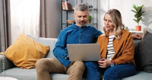 Caucasian middle-aged happy married couple bearded man and beautiful woman sitting on couch in good mood typing on laptop browsing online searching internet at home in cozy room, leisure concept photo