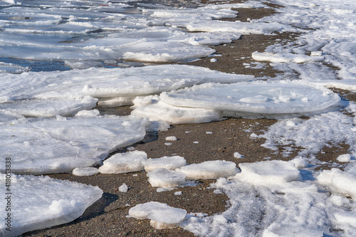 Spring background with melting ice on the surface of a frozen bay. Snowy shore of a frozen lake.