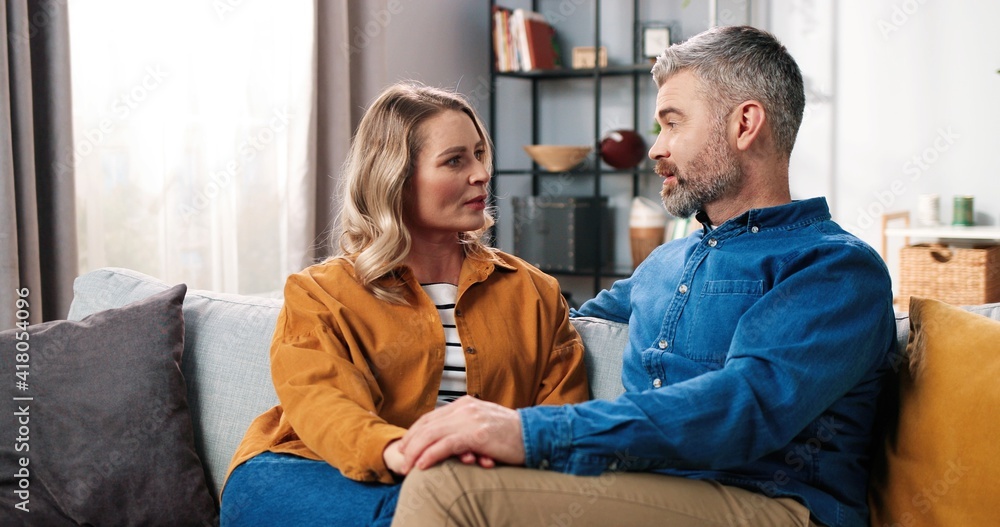Portrait of cheerful happy Caucasian adult middle-aged couple wife and husband chatting having nice conversation while resting at home sitting on couch in positive mood, love family concept Portrait of cheerful happy Caucasian adult middle-aged couple wife and husband chatting having nice conversation while resting at home sitting on couch in positive mood, love family concept