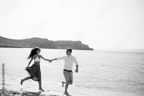 Happy cheerful couple hold hans and running in the sea together with splashes of water on a tropical beach at sunset. Romantic vacation, honeymoon love